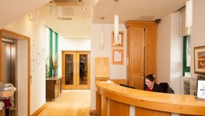 A reception area with a wooden reception desk, a counter, and a person seated behind it. There is a hallway with wooden flooring and a door visible in the background. - The Arches Hotel (Galway)