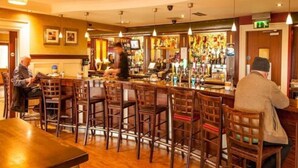 A bar area with wooden chairs and tables, a counter with various bottles, and a television screen. - The Arches Hotel (Galway)
