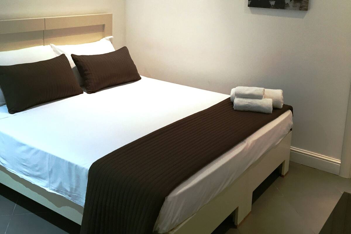 Una cama bien tendida con ropa de cama blanca y marrón, cabecera y dos cuadros colgados en la pared.