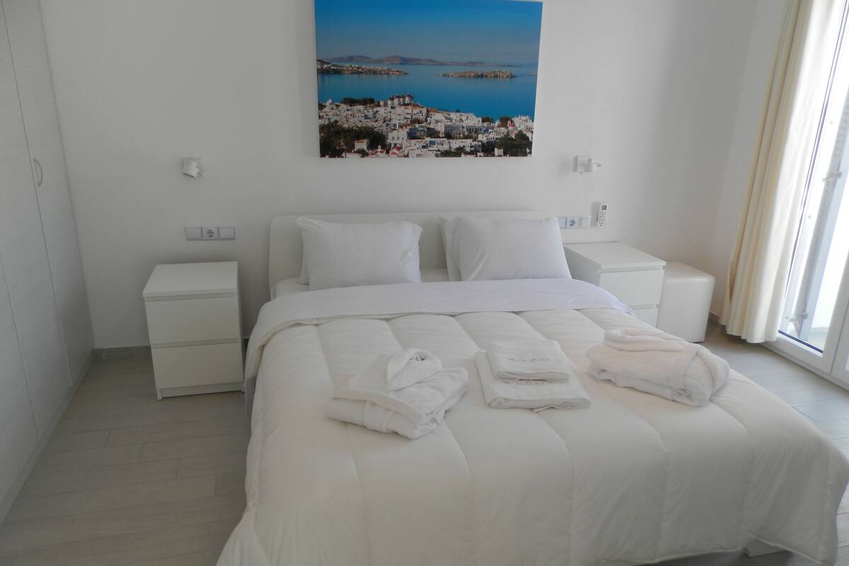 Ein Hotelzimmer mit einem großen Bett, weißer Bettwäsche, einem Nachttisch und einem Bild einer Küstenlandschaft.