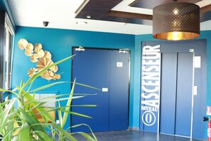 A blue elevator with 'ASCENSEUR' and 'NIVEAU' signage, a modern light fixture, and a plant in the foreground. - Hôtel Orly Superior (Athis-Mons)