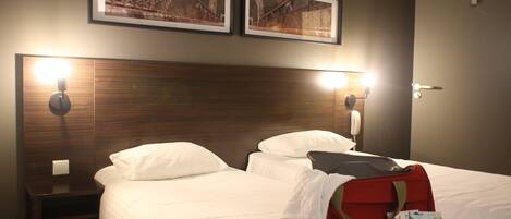 Habitación de hotel con dos camas, cabeceros de madera, luces fijadas a la pared y cuadros enmarcados sobre las camas.