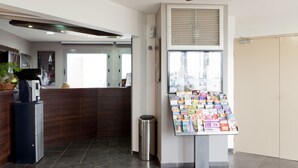 A reception area with a wooden reception desk, a coffee machine, a trash bin, and a display of magazines. - Hôtel Orly Superior (Athis-Mons)
