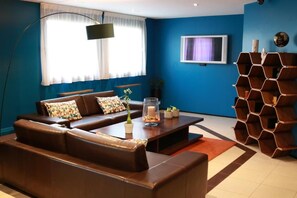 A modern living room with a brown leather sofa, a coffee table with decorative items, a TV on the wall, and a unique hexagonal wooden shelf. - Hôtel Orly Superior (Athis-Mons)
