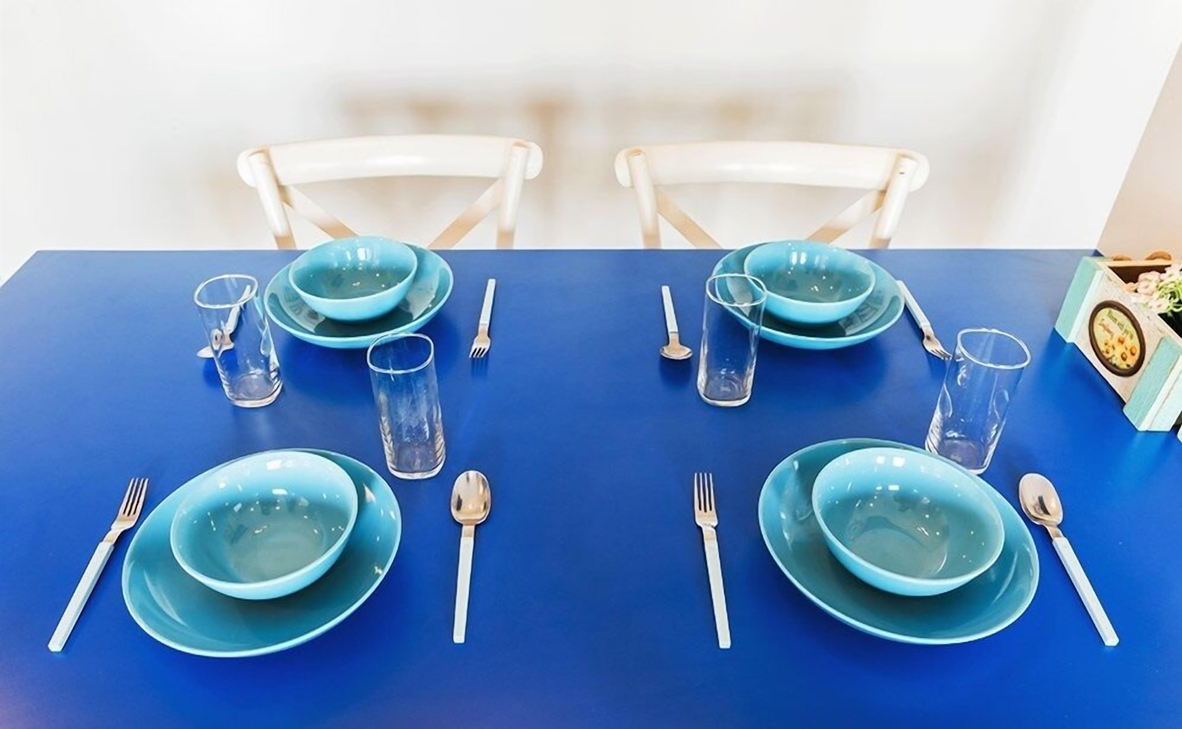 A dining table set with blue plates, bowls, and cutlery, surrounded by white chairs.