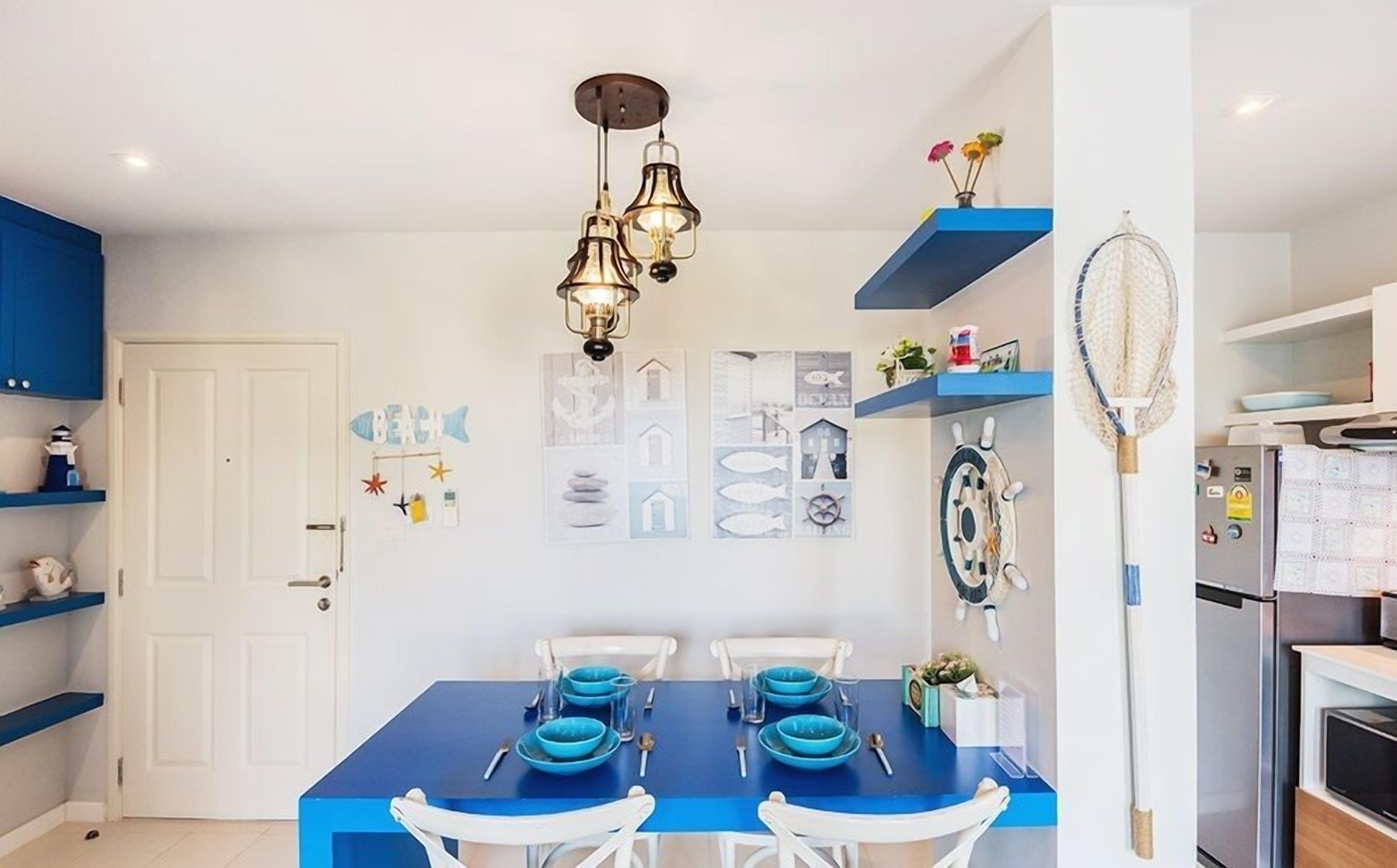 Comedor con una mesa azul dispuesta para dos, sillas blancas, una lámpara de pared y decoración temática náutica.