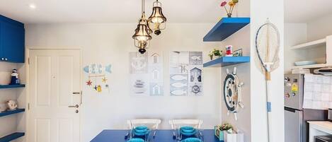 Comedor con una mesa azul dispuesta para dos, sillas blancas, una lámpara de pared y decoración temática náutica.