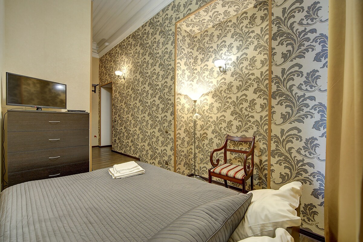 A hotel room with a bed, a TV on a dresser, a chair, and patterned wallpaper.