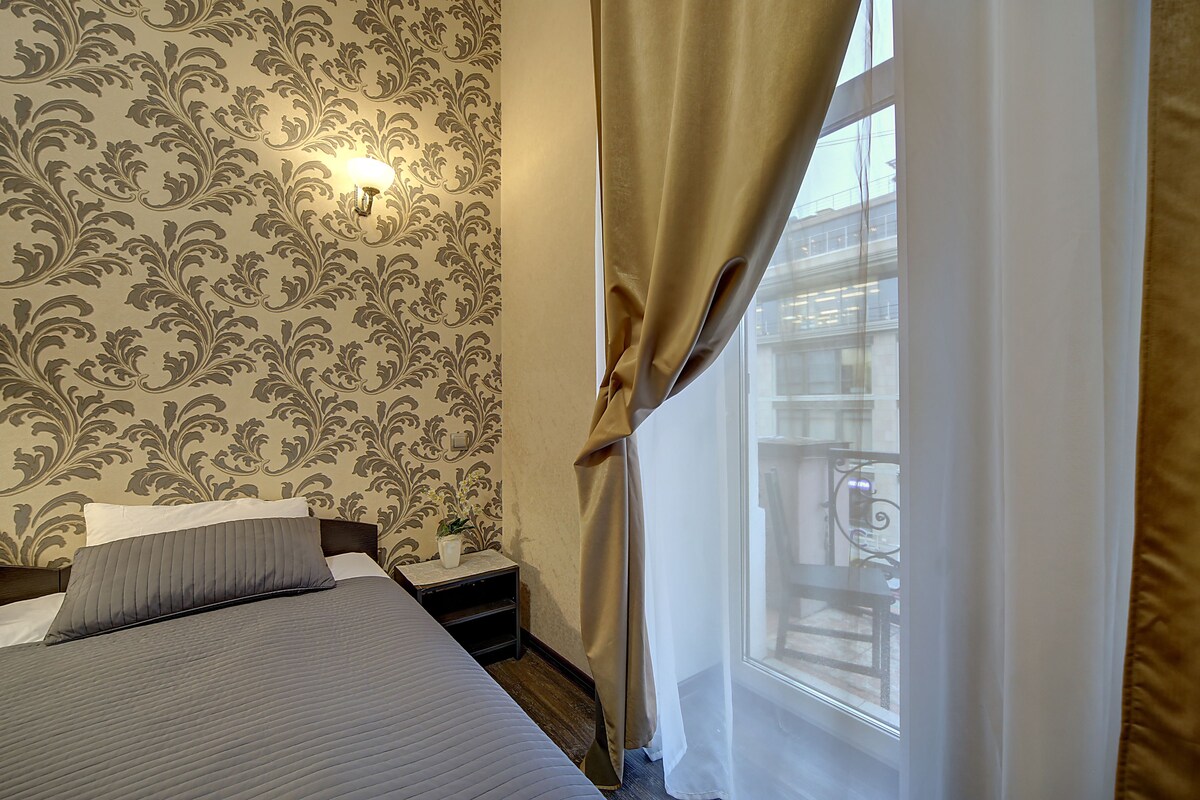 A bedroom with a bed, a nightstand, a potted plant, and a window with curtains.