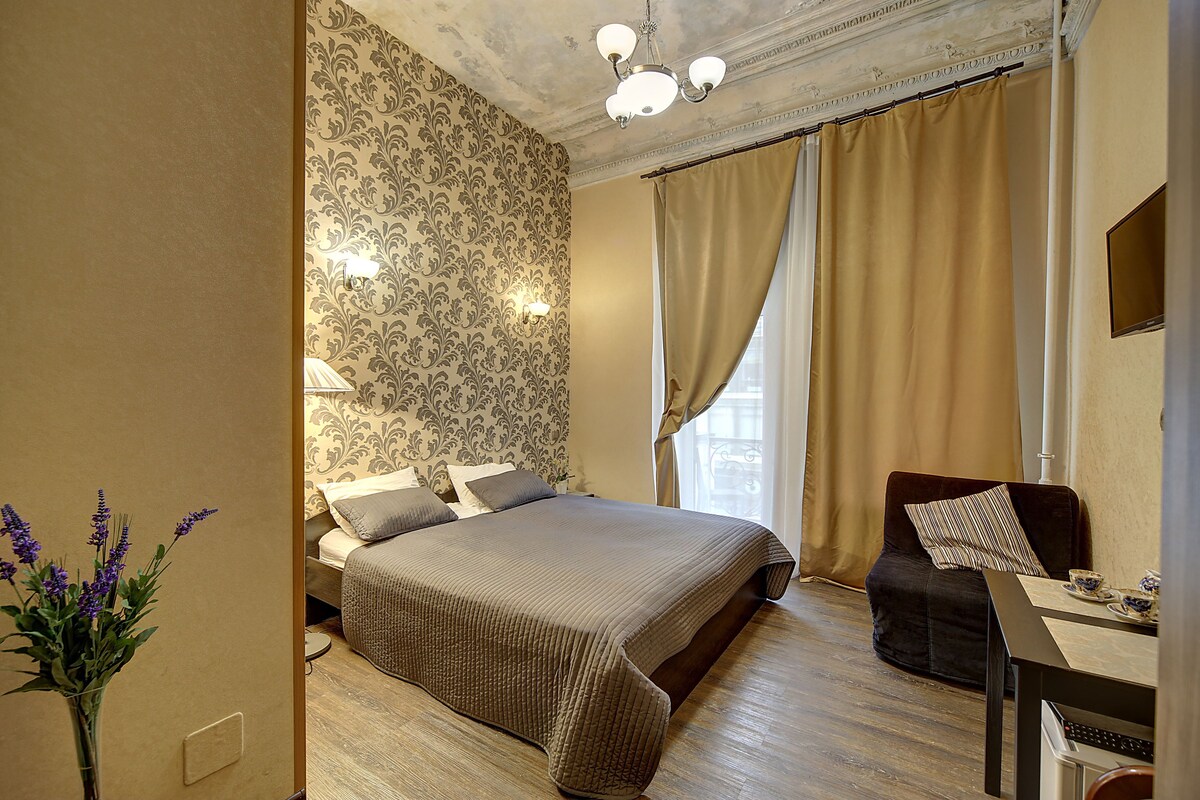 A hotel room with a large bed, a sitting area with a chair and table, and a vase with flowers.