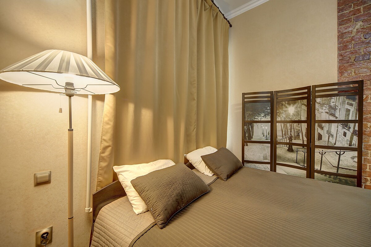 A bedroom with a bed, a lamp, a window with a view of a building, and a curtain.