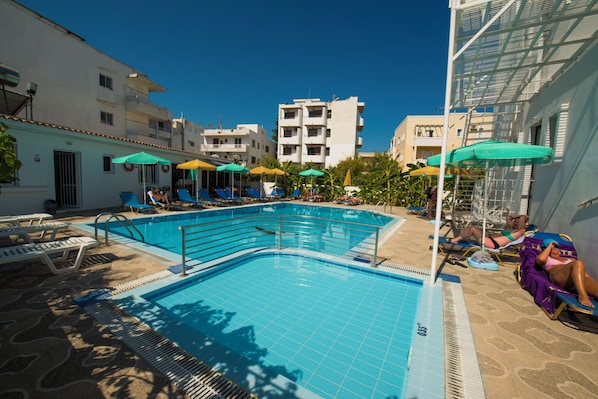Une piscine entourée de bâtiments, avec des chaises longues et des parasols.