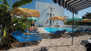 Area piscina con poltrone blu, ombrelloni e un edificio bianco sullo sfondo.