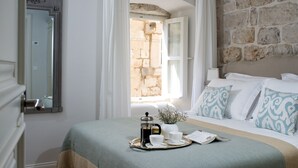 Une chambre à coucher bien rangée, avec un lit, une table de chevet sur laquelle se trouve un plateau avec des articles pour le petit-déjeuner, une fenêtre avec des rideaux et un mur en pierre.
