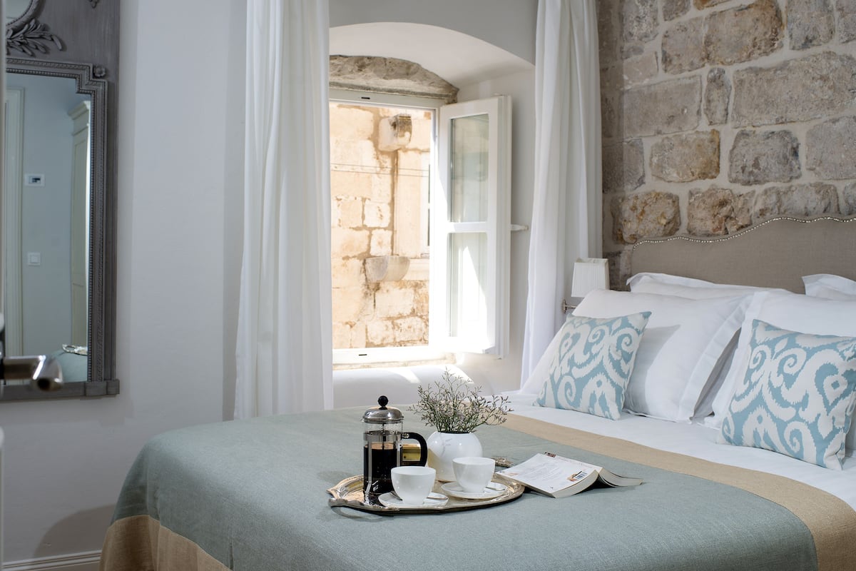 Un dormitorio ordenado con una cama, una mesita de noche con una bandeja de desayuno, una ventana con cortinas y una pared de piedra.