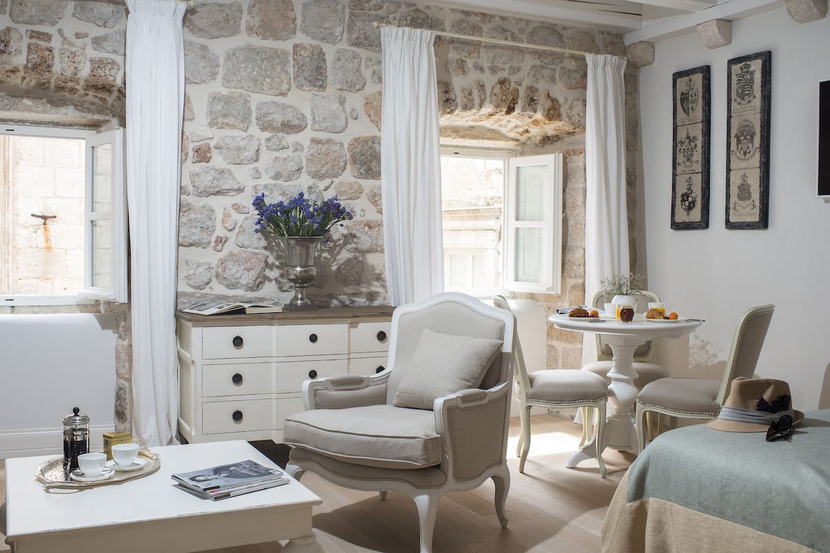 Una habitación con paredes de piedra, una silla blanca, una mesa con una bandeja y una cama con un sombrero encima.