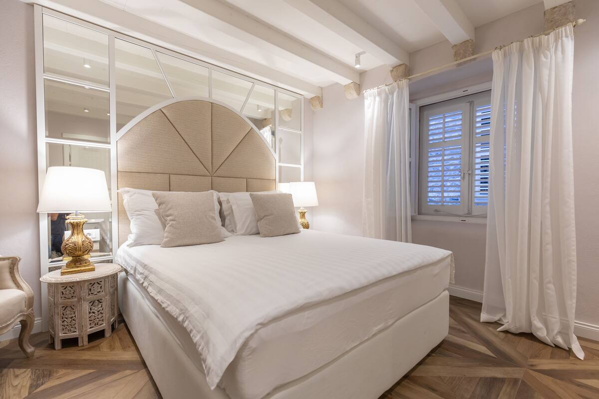 Un dormitorio con una cama grande, una mesita de noche con lámpara, un espejo y una ventana con cortinas.