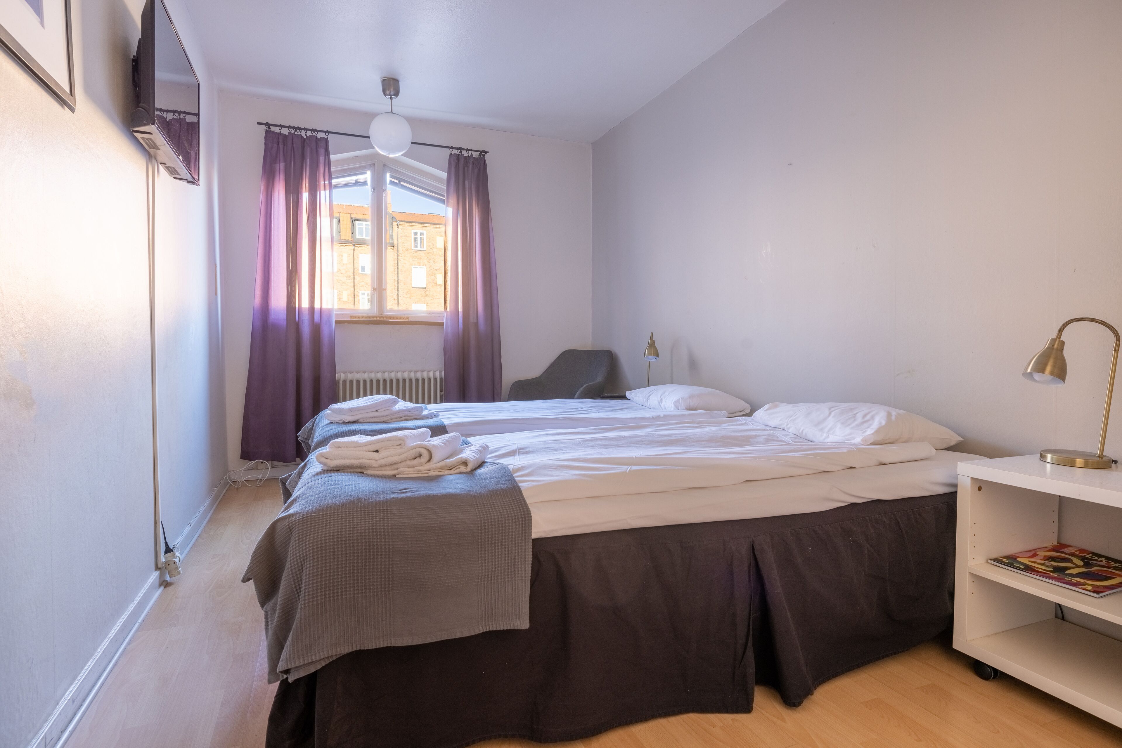 Una cama individual con colcha gris, una lámpara de noche y una ventana con cortinas moradas.