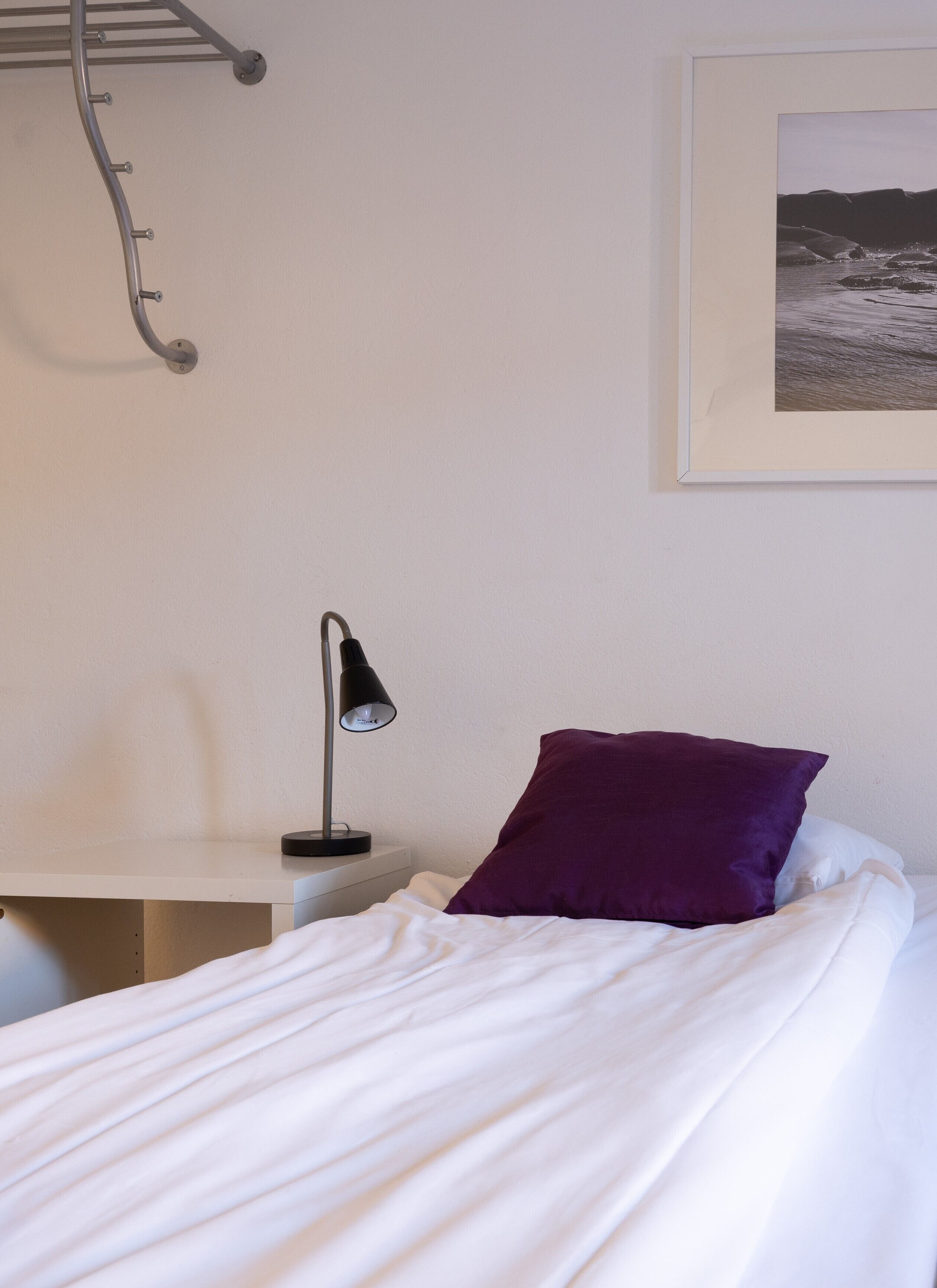 Un dormitorio con cama, mesita de noche, lámpara, almohada morada y un cuadro en la pared.