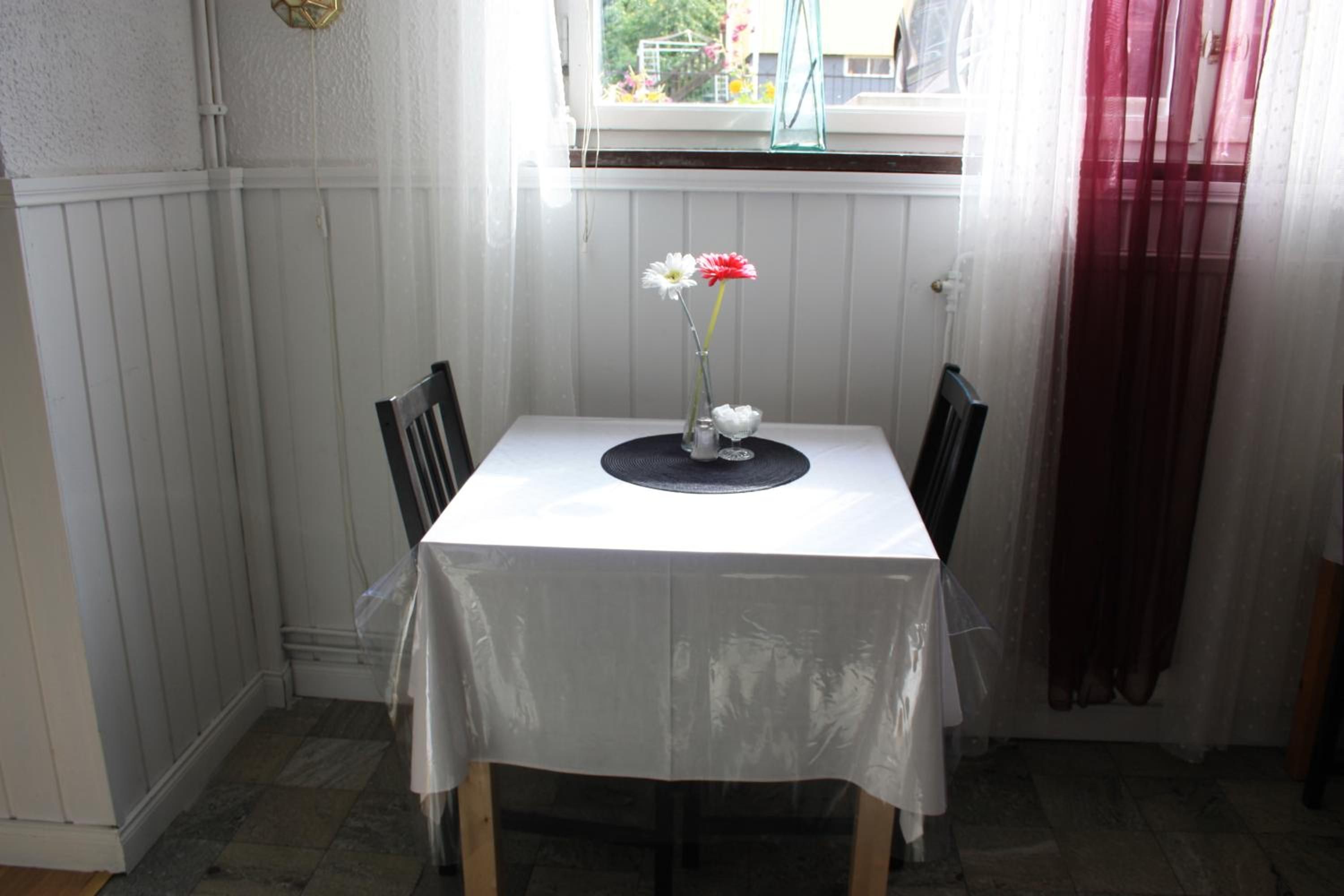 Ein kleiner Esstisch mit weißer Tischdecke, zwei Stühle, eine Blumenvase und ein Fenster mit Blick auf grüne Landschaft.