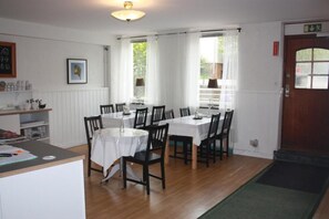 A dining area with a wooden floor, white tables and chairs, and a view of the outdoors through large windows. - Lilla Hotellet (Eskilstuna)
