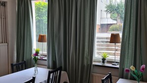 A dining area with a table, chairs, and a window with curtains. There are potted plants on the windowsill. - Lilla Hotellet (Eskilstuna)