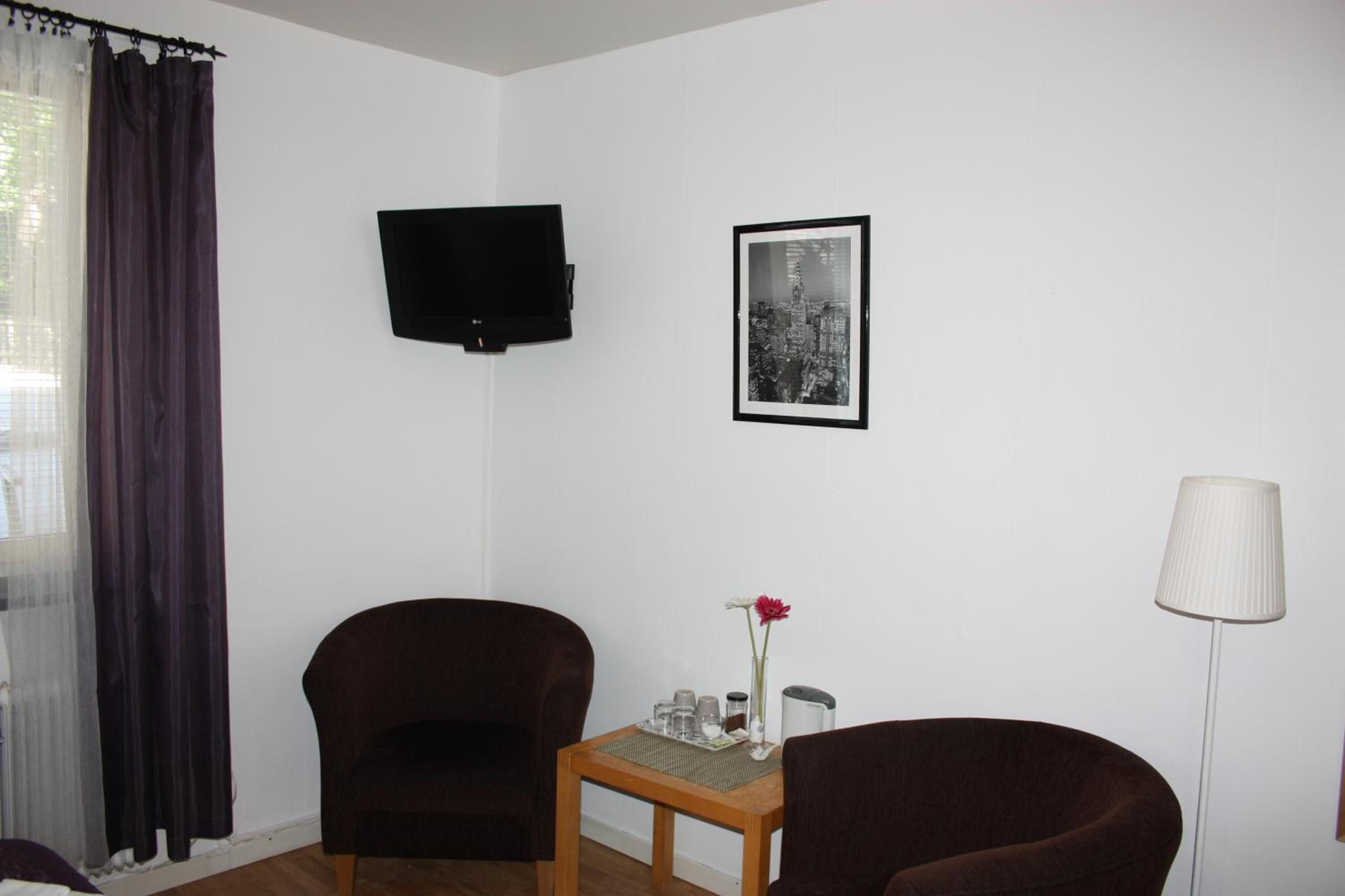 A room with two dark armchairs, a small wooden table with a vase and glasses, a framed black and white cityscape photo, a flat-screen TV, and a lamp.