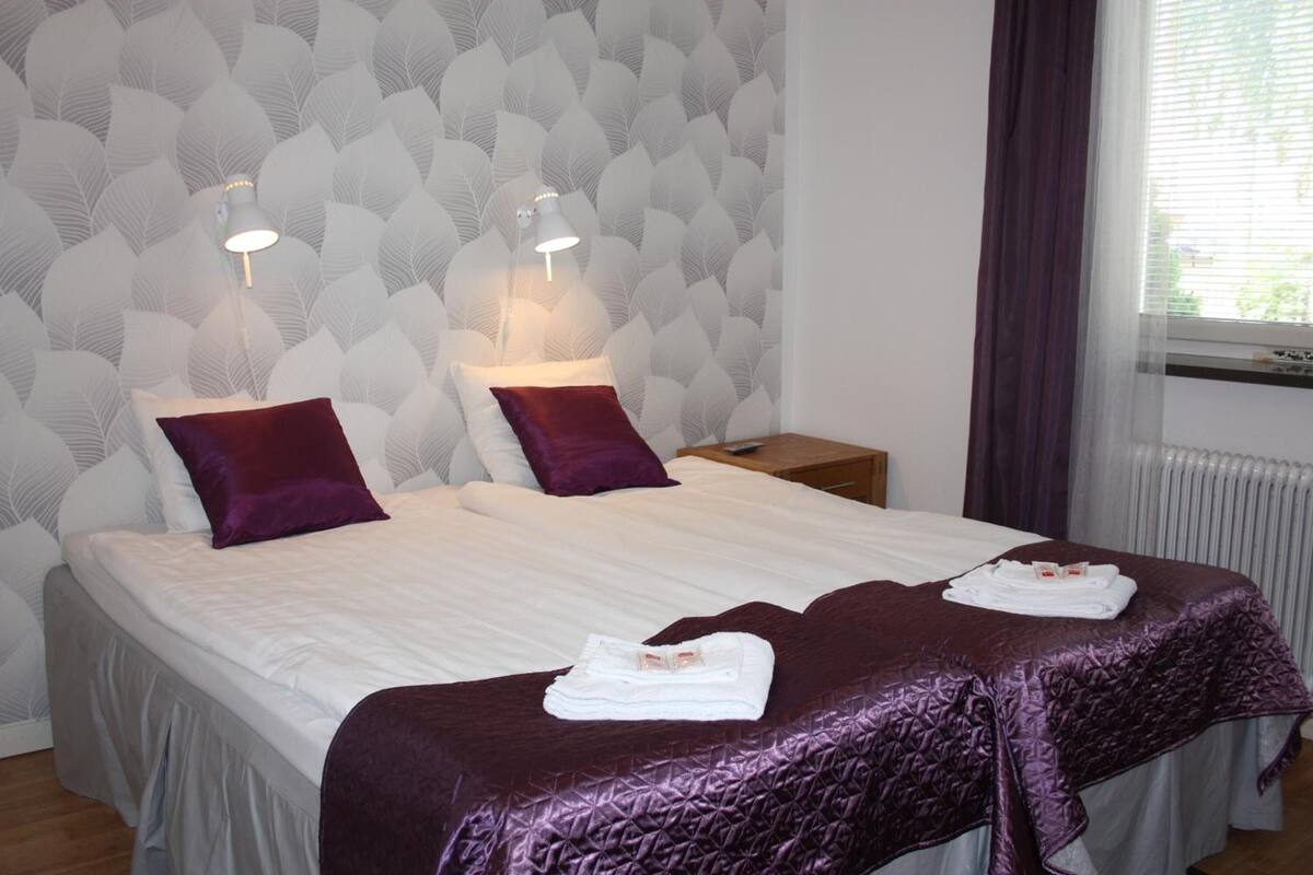 A double bed with a white and purple bedspread, two purple pillows, and a folded towel on the bed. There is a nightstand with a lamp and a wooden side table with a remote control. The room has a patterned wallpaper and a window with curtains.