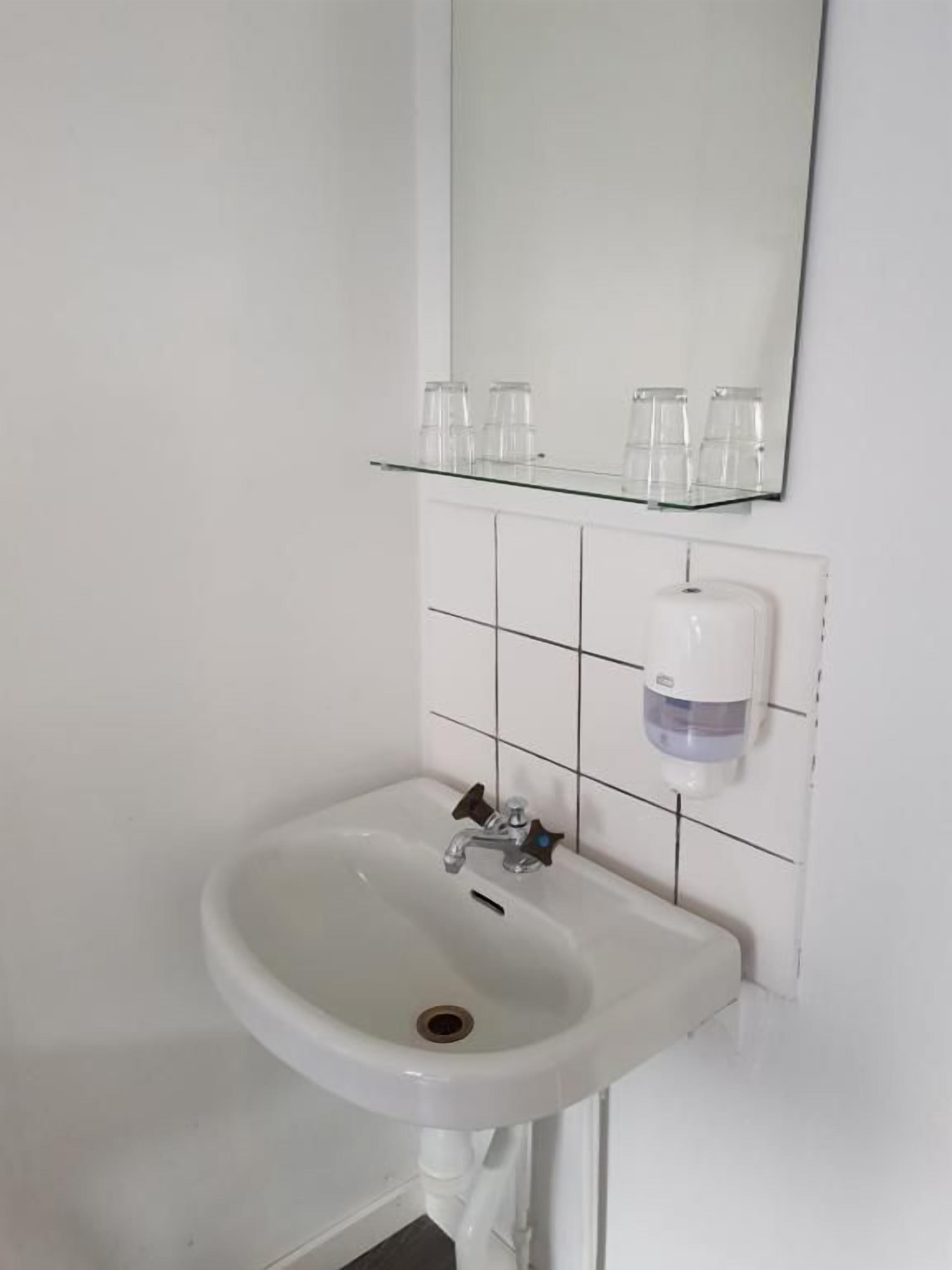 Een witte badkamer met een glazen plank waarop drie glazen staan, een witte wastafel met een kraan en een zeepdispenser aan de muur.