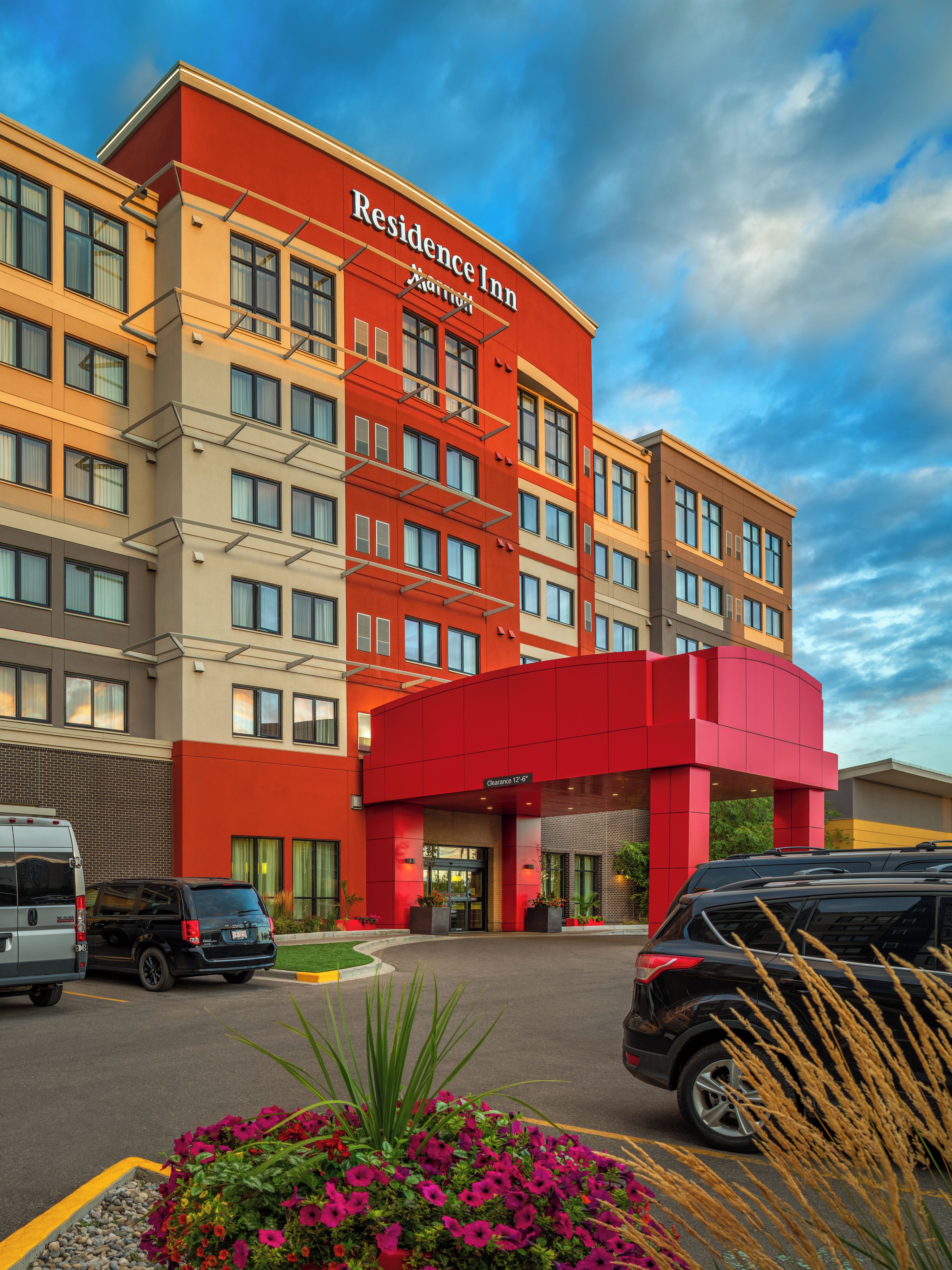 A multi-story hotel with a red and beige facade, a clear sky, and a parking lot with vehicles.