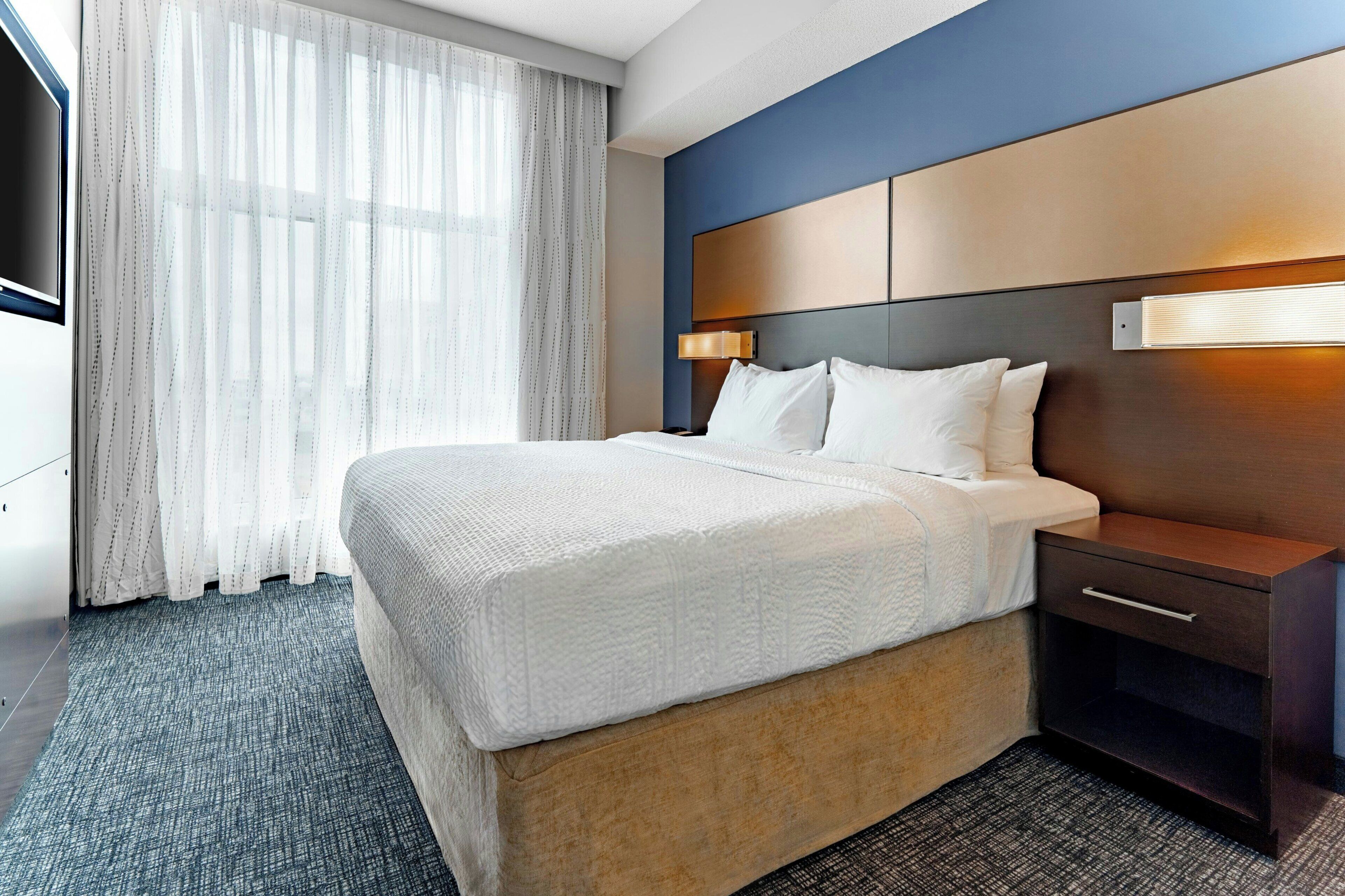A hotel room with a bed, bedside table, lamp, and a large window with curtains.