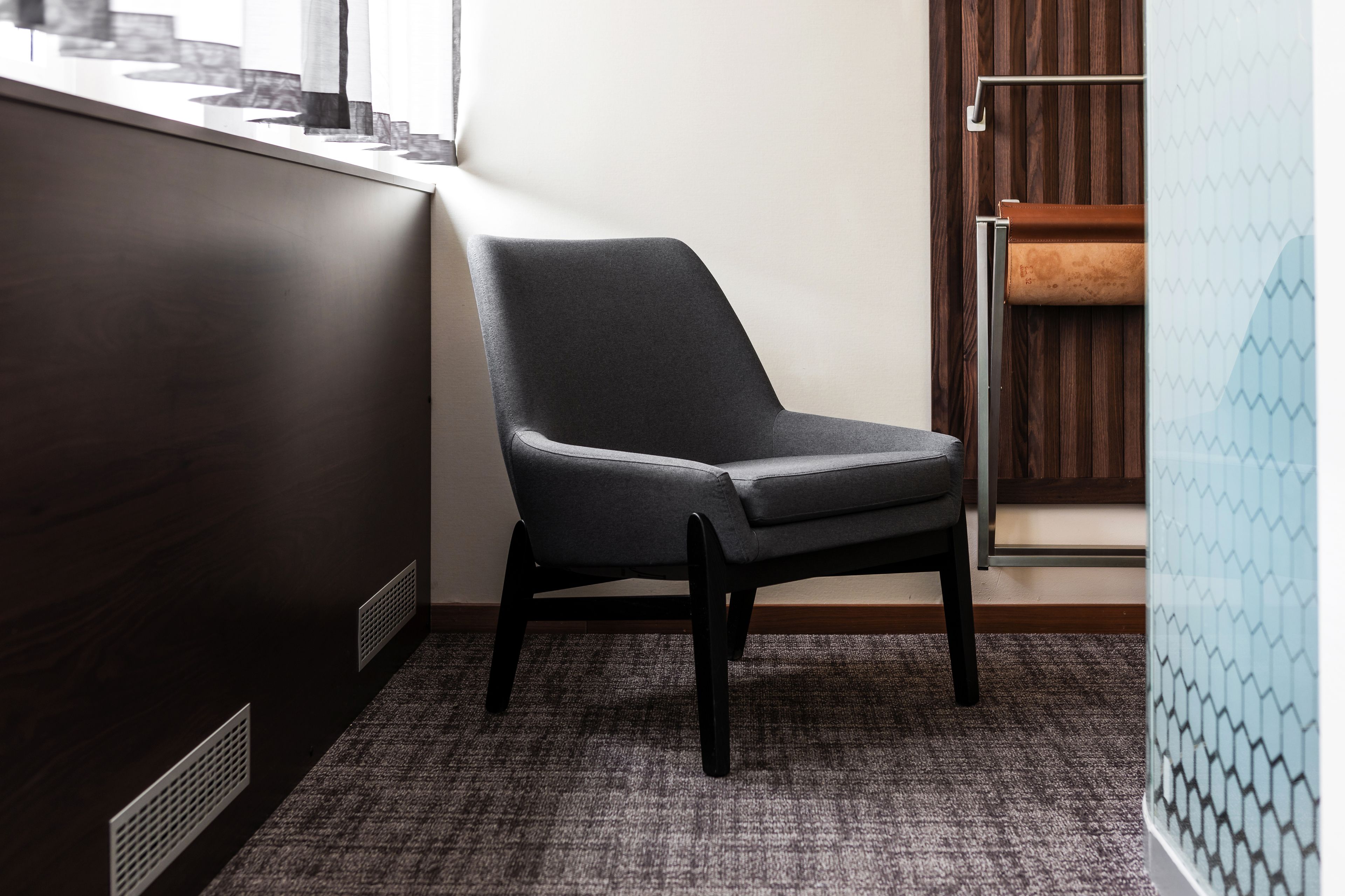 A corner of a hotel room with a dark wooden floor, a grey upholstered chair, and a patterned wall.