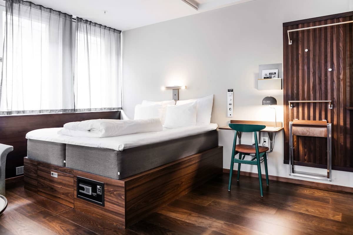 A modern hotel room with a bed, a desk, a chair, and a wardrobe.