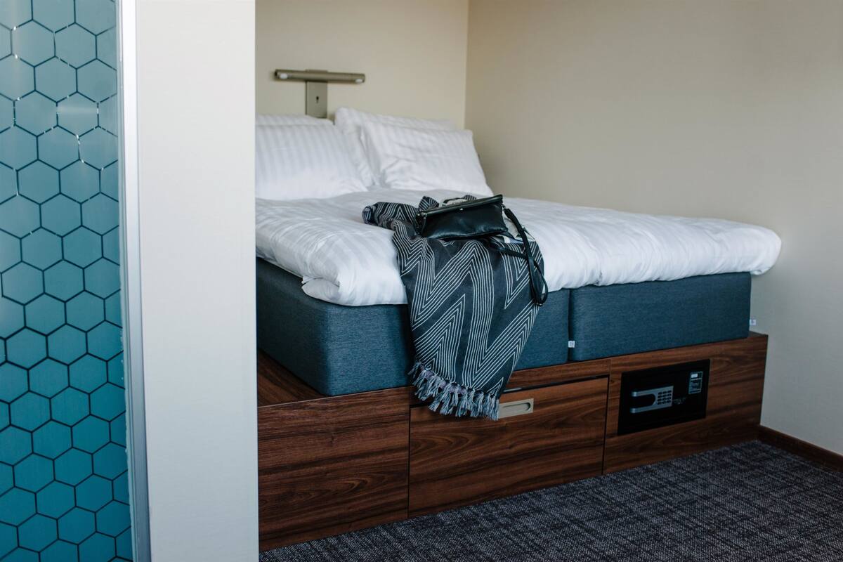 A neatly made bed with a white comforter and pillows, a blue blanket, and a black bag on the side. There is a wooden drawer unit with a microwave below the bed.