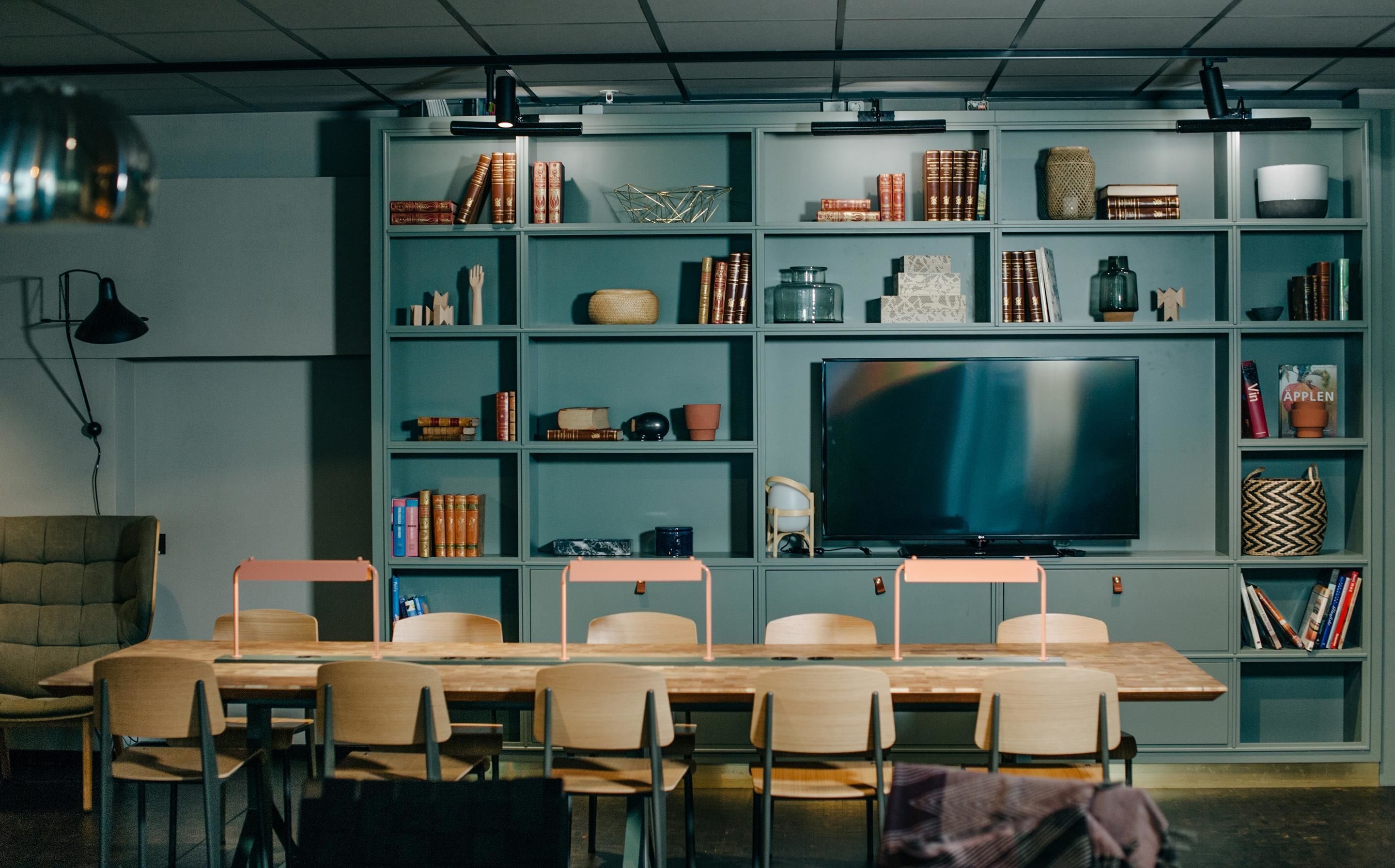 Une salle de réunion moderne équipée d’un grand téléviseur à écran plat, d’une table en bois avec des chaises et d’une bibliothèque remplie de livres et d’objets décoratifs.