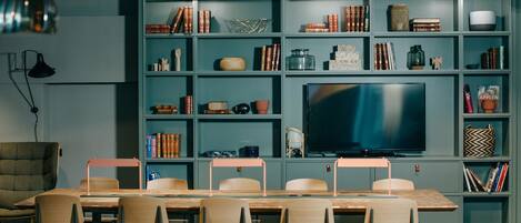 Una sala de reuniones moderna con una gran televisión de pantalla plana, una mesa de madera con sillas y una estantería llena de libros y objetos decorativos.