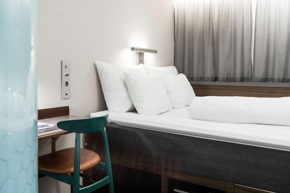 Una habitación de hotel con una cama, un escritorio con una silla, un enchufe y una ventana con cortinas.