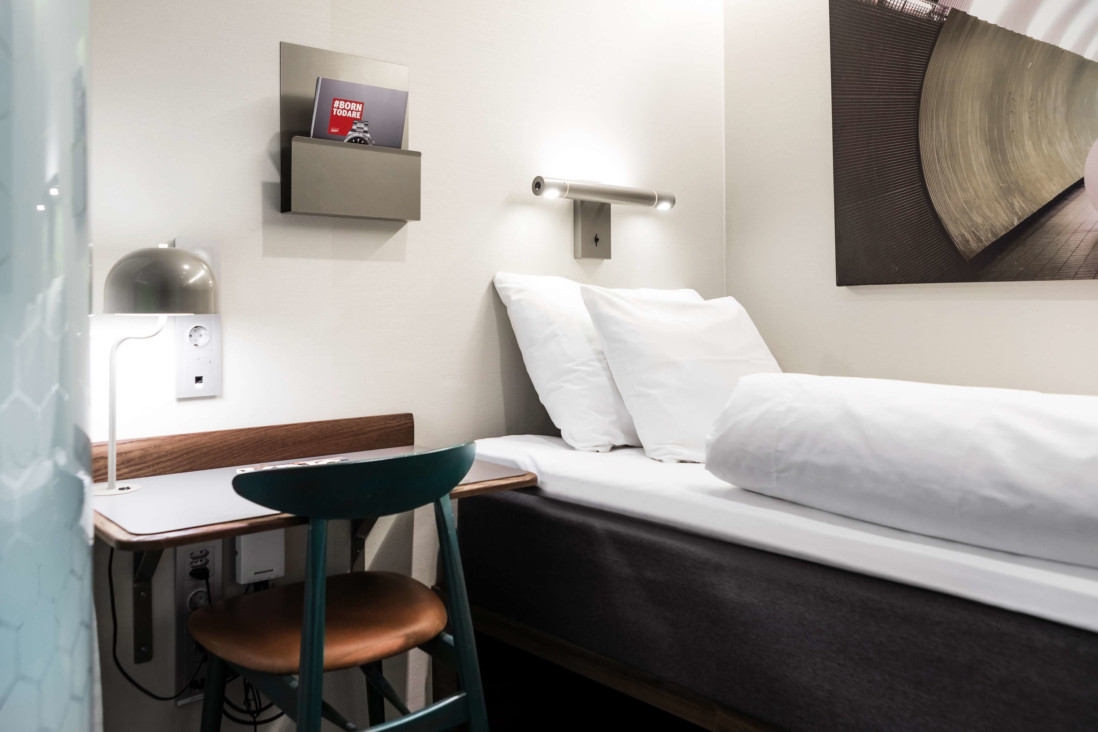 Une chambre d’hôtel comprenant un lit, un bureau avec une chaise, une étagère avec un livre et un luminaire fixé au mur.