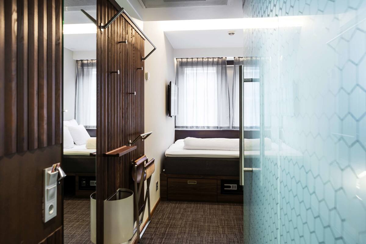 A compact hotel room with a bed, a small desk, a chair, a mirror, and a window with curtains.
