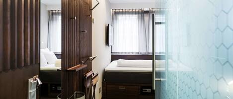 Quarto de hotel pequeno e funcional, com uma cama, uma pequena escrivaninha, uma cadeira, um espelho e uma janela com cortinas.