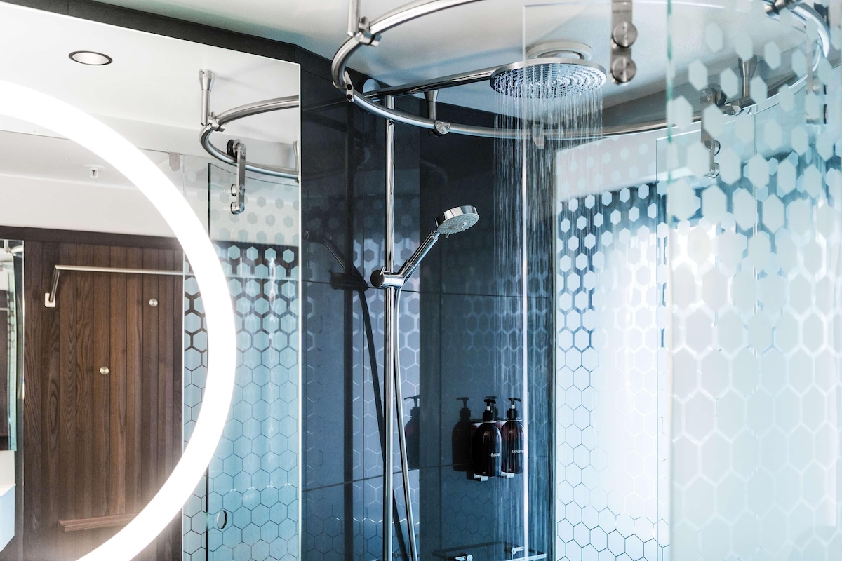 Ducha moderna con cabina de vidrio, puerta de madera y una lámpara circular.