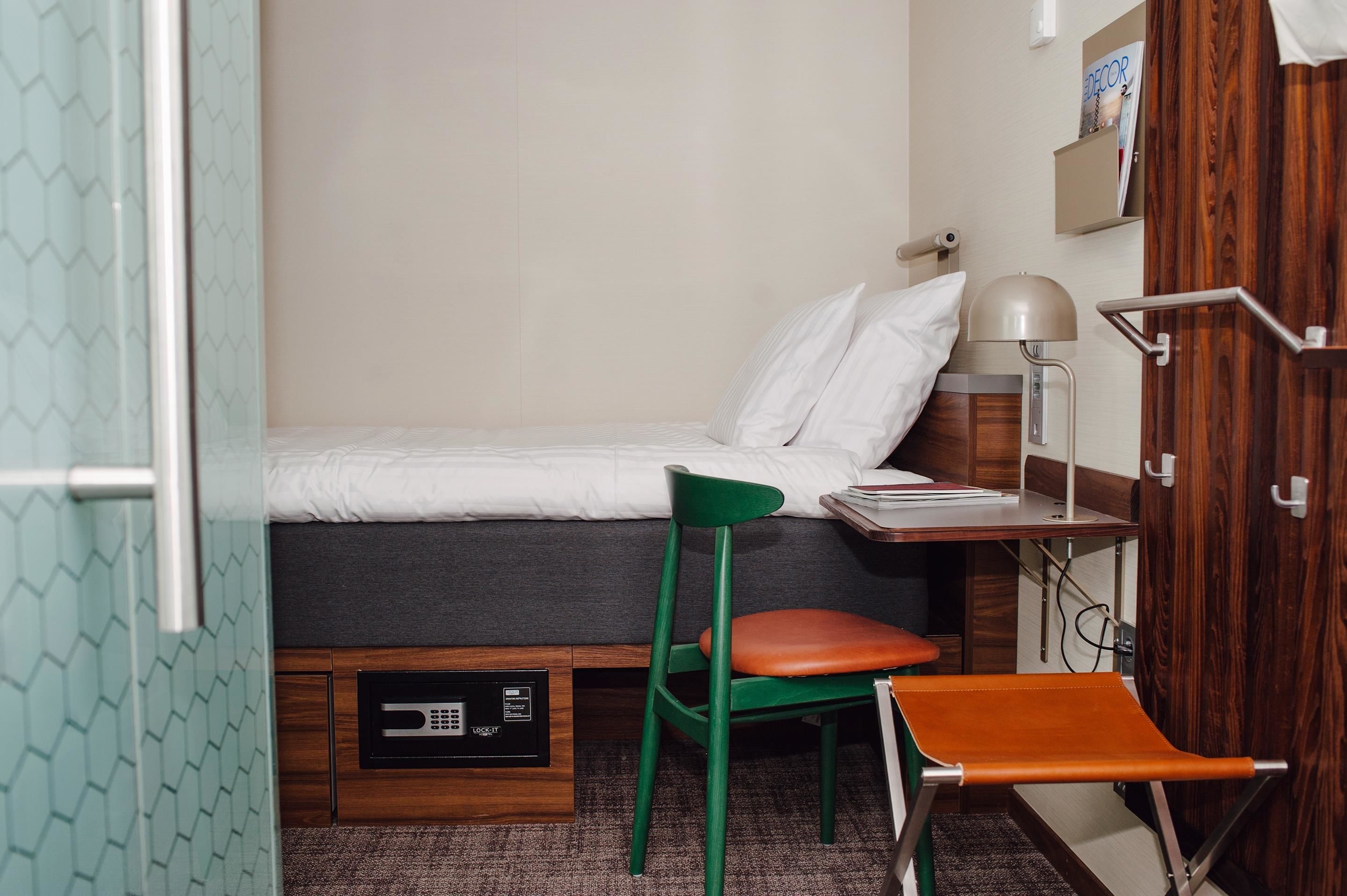 Una habitación de hotel pequeña con una cama, una silla verde, una mesita con lámpara y un armario de madera.