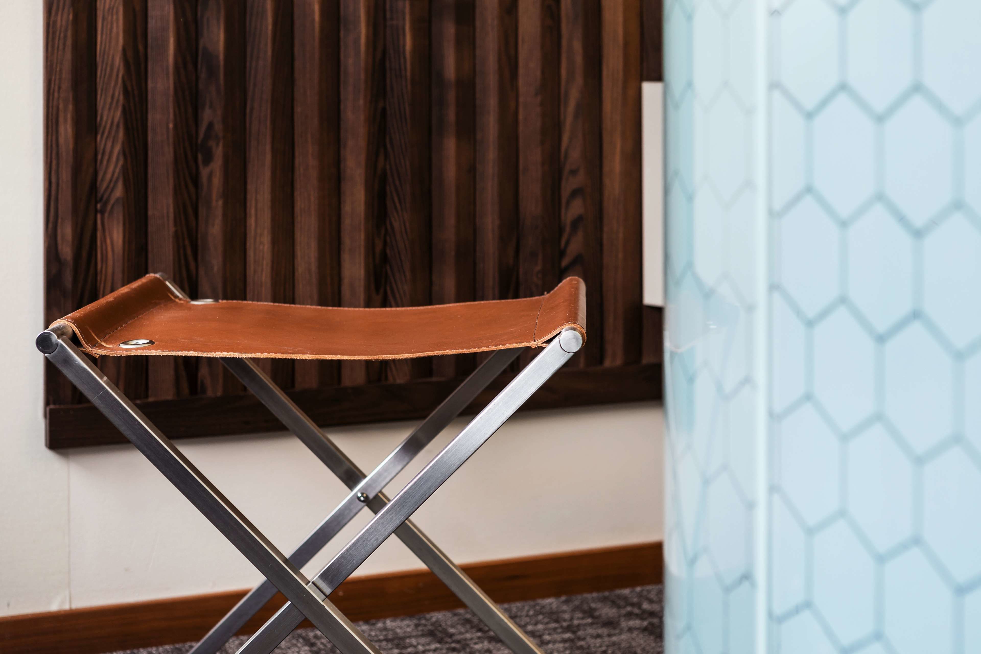 Una silla plegable de cuero marrón con patas metálicas, ubicada en una habitación con panel de madera y pared azul claro con un diseño hexagonal.