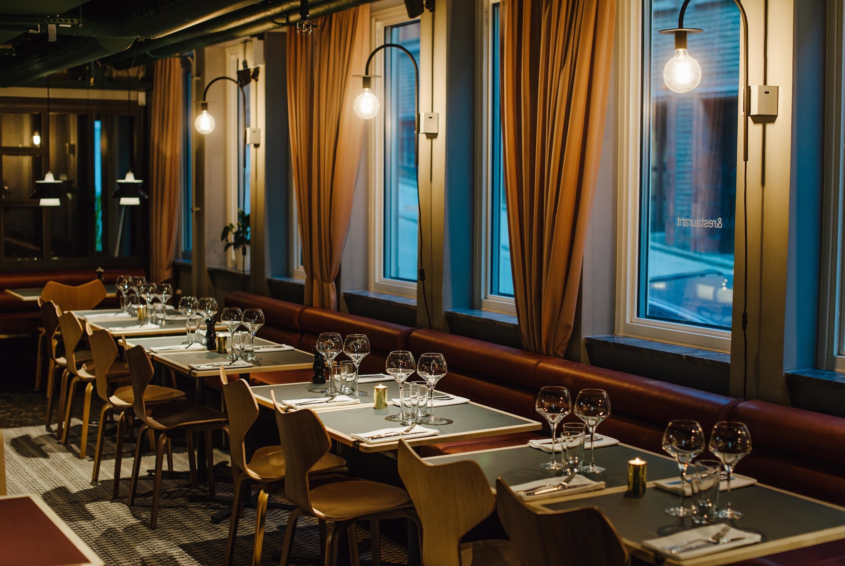 A restaurant with tables set for dining, a long bench along the wall, and large windows with curtains.