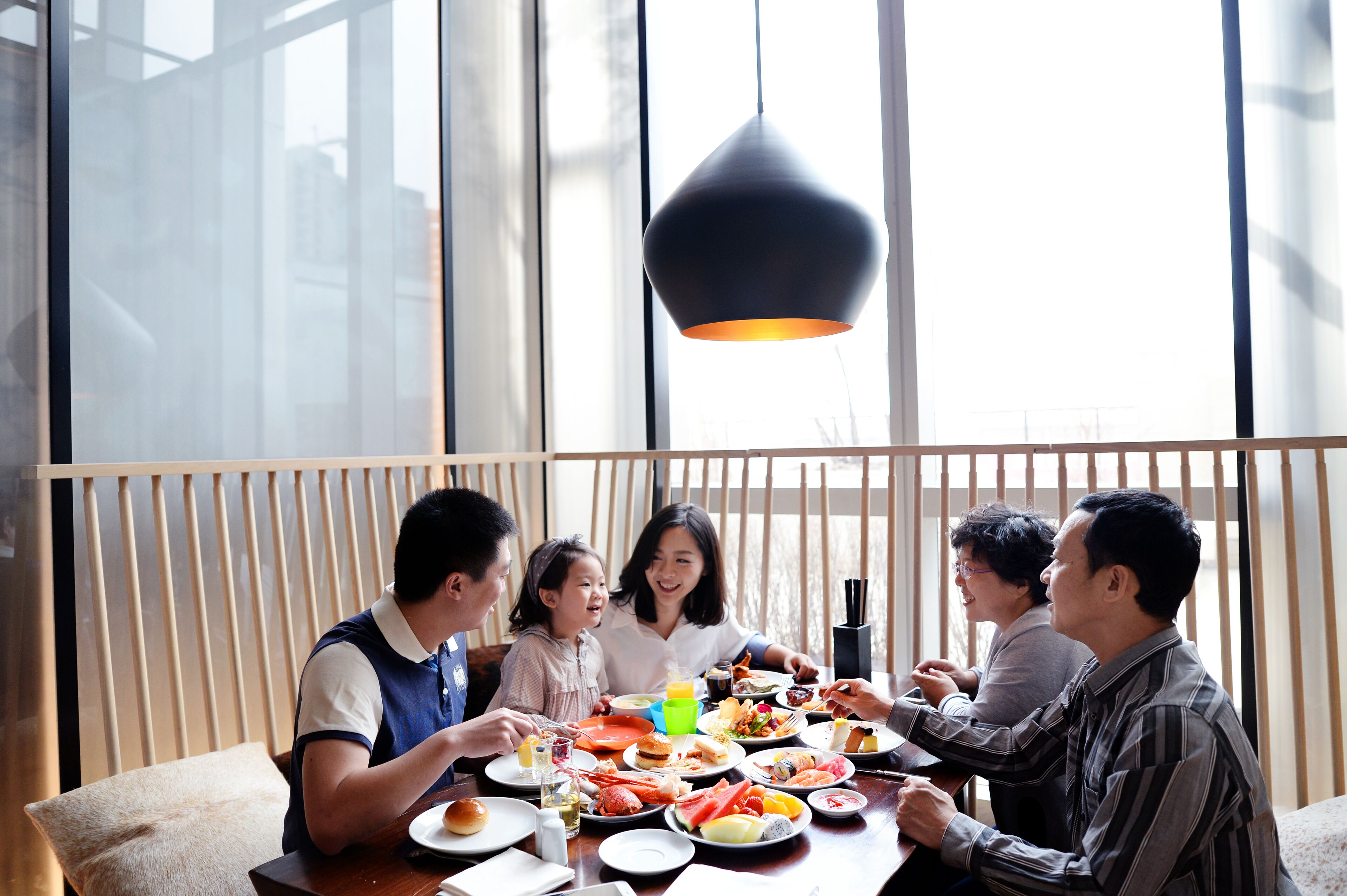 Un grupo de personas cenando en una mesa con diversos platos, en una habitación con ventanas amplias y una lámpara colgante.