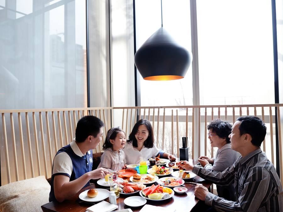 A group of people dining at a table with various dishes, in a room with large windows and a hanging light fixture.