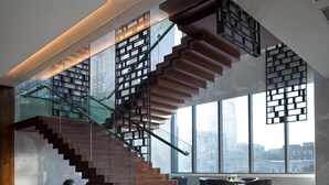 Un escalier moderne avec des rampes en verre et des marches en bois, menant à un étage supérieur doté de grandes fenêtres offrant une vue sur la ville.