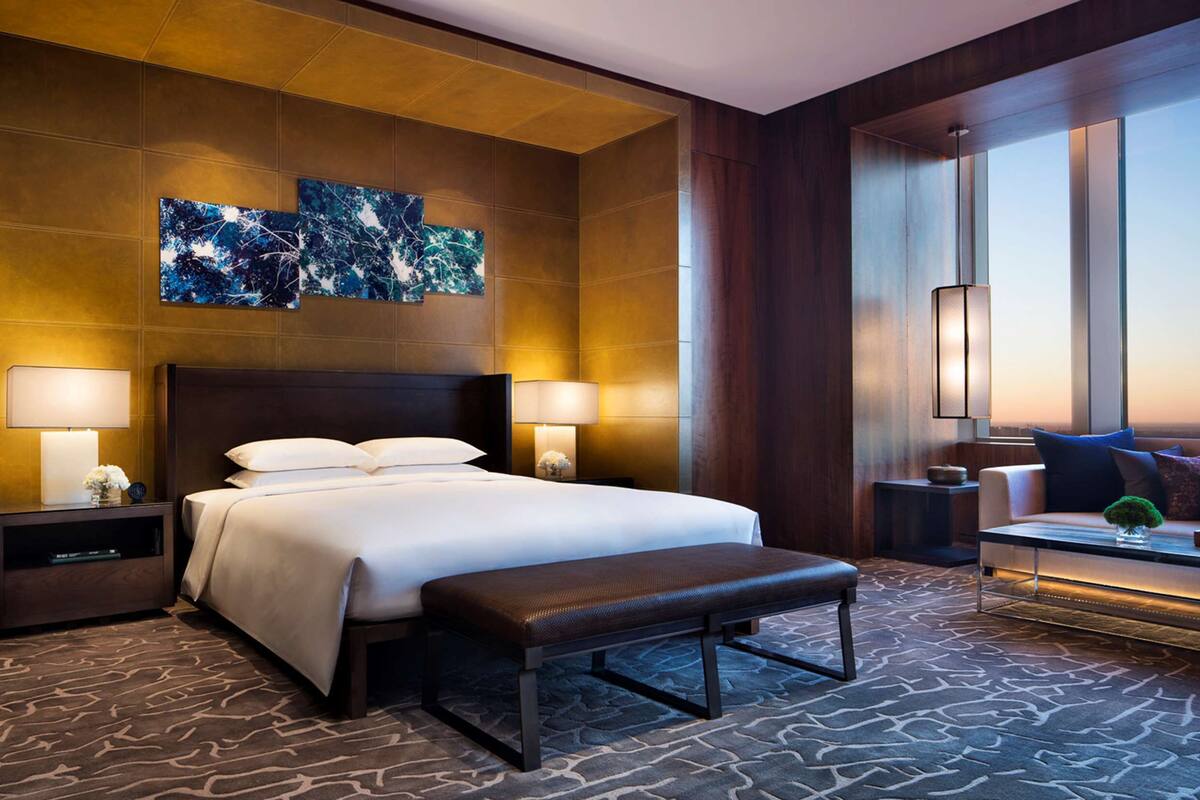 Habitación de hotel con una cama grande, dos mesitas de noche con lámparas, un banco, un ventanal amplio y una obra de arte abstracto en la pared.