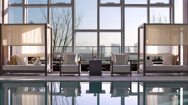 Un espace piscine intérieur moderne, doté de deux coins salon, chacun comprenant un canapé et une table, ainsi qu’une grande fenêtre offrant une vue sur le panorama urbain.