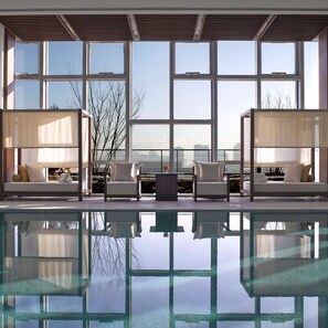 Un espace piscine intérieur moderne, doté de deux coins salon, chacun comprenant un canapé et une table, ainsi qu’une grande fenêtre offrant une vue sur le panorama urbain.